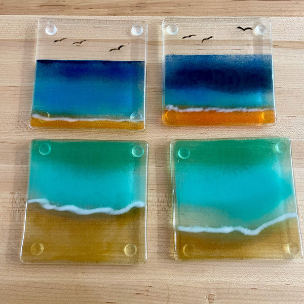 Load image into Gallery viewer, A set of four fused glass coasters in various shades of blue and aqua featuring a beach scene, arranged on a maple wood table background