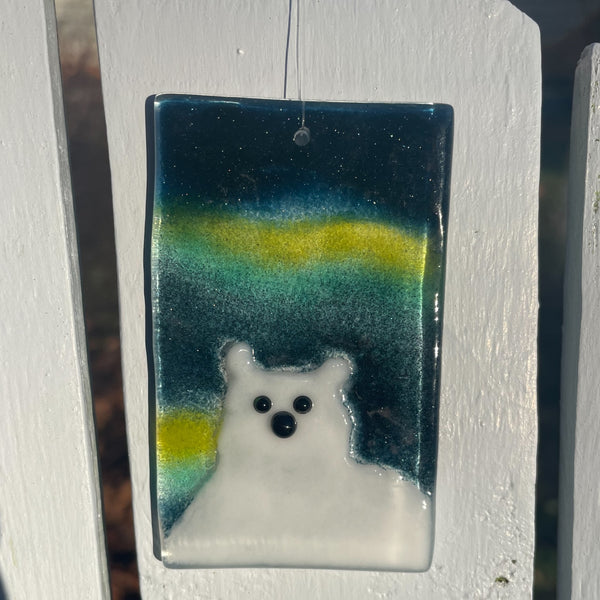 Load image into Gallery viewer, A glass tile featuring a white polar bear in front of the aurora. The tile is balanced on a pale blue Adirondack chair.