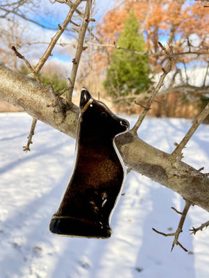 A black glass shepherd dog ornament hangs from a tree. It's a snowy scene. This dog is Belgian Shepherd Dog in a series of Dog designs.