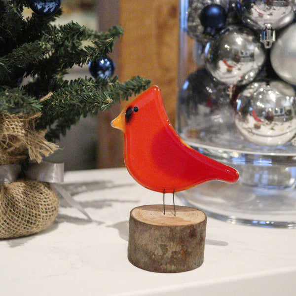 Load image into Gallery viewer, A fused glass red cardinal adult bird perched on a log slice, with christmas decorations in the background.