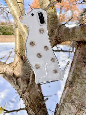 A white and black spotted glass Dalmation dog hangs from a tree. It's a snowy scene. This dog is part of a series of Hanging Glass Dog designs.