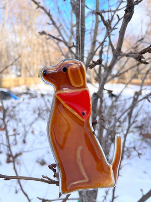 A brown glass dog wearing a red glass bandana hangs from a tree. It's a snowy scene. This dog is Dog B of a series of Dog designs.
