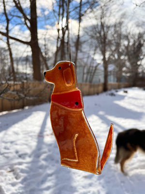 A chestnut brown glass dog wearing a red glass bandana hangs from a tree. It's a snowy scene with the rear of a Rough Collie in the background. This dog is Dog B of a series of Dog designs.