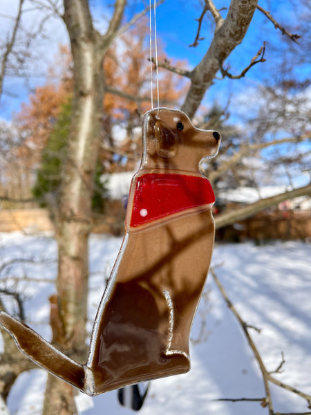 Load image into Gallery viewer, A mid brown glass dog wearing a red glass bandana hangs from a tree. It's a snowy scene. This dog is Dog D of a series of Dog designs.
