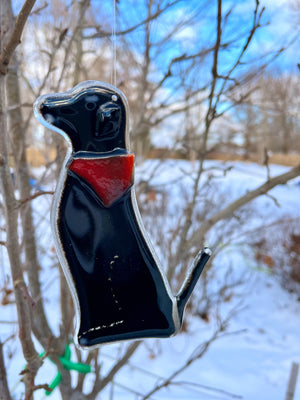A black glass dog wearing a red glass bandana hangs from a tree. It's a snowy scene. This dog is Dog E of a series of Dog designs.