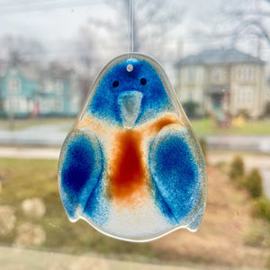 A blue and chestnut brown glass bluebird hanging ornament hangs from a window. In the background are old townhouses.