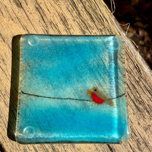 A blue fused glass coaster featuring a robin on a wire. The coaster is laying on a rustic yellow piece of wood.