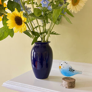 Navy blue vase with sunflowers and other flowers on a white surface with a yellow wall background. Beside the vase of flowers is a fused glass budgie made of blue and white glass. It's perched on a log slice.
