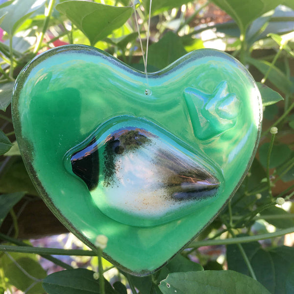 Load image into Gallery viewer, A green fused glass heart featuring a chickadee bird hangs in front of plants