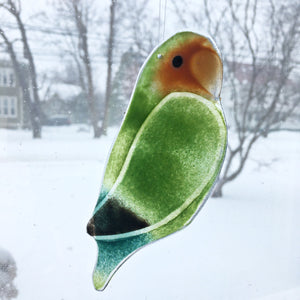 A green fused glass lovebird parrot hangs in a window. In the background is a snowy scene and Georgian townhous