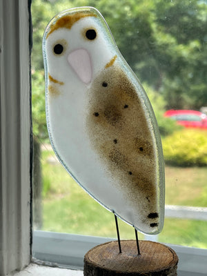 Decorative owl ornament on a wooden base in front of a window with a blurred outdoor view.