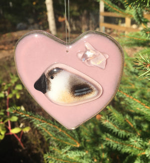 A Fused Glass Pink Heart with Chickadee hangs in front of a pine tree
