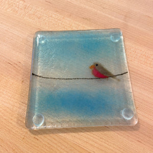 Square glass coaster with a fused glass bird on a wire design, placed on a wooden surface.