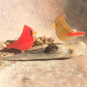 Two fused glass cardinals, one red male and one an amber female, are perched on driftwood. The background is a weathered metal.