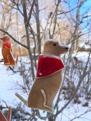 A Glass Brown Hanging Dog with Bandana dangles from a bare tree branch. In the background is a snowy scene another hanging dog ornament.
