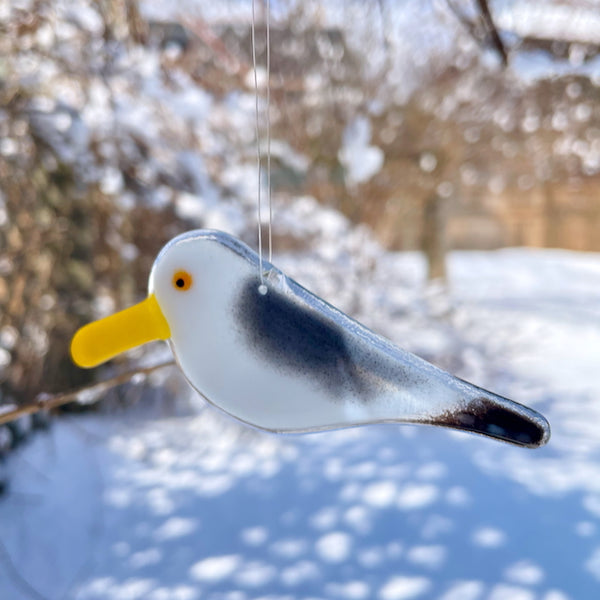 Load image into Gallery viewer, grey, white and black fused glass seagull chick window hanging ornament, against background of a snowy garden yard