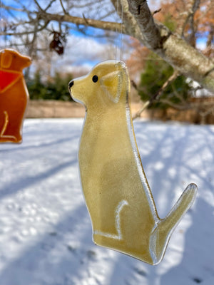 A brown glass dog in the shape of a pale Labrador Retriever hangs from a tree. It's a snowy scene. This one of a series of Dog designs.