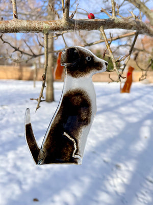A black and white glass dog in the form of a border collie bandana hangs from a tree. It's a snowy scene. This dog is Dog B of a series of Dog designs.