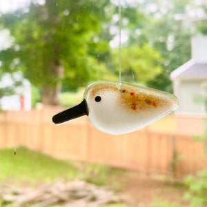 A brown and white speckled fused glass pocket sandpiper hangs in a window