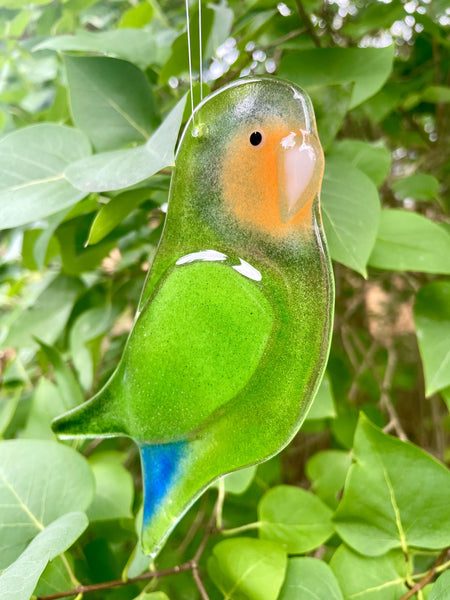 Charger l'image dans la galerie, A green parrot-shaped glass hanging ornament with an orange beak, white eye patch, and blue tail accent, suspended against a background of green leaves.