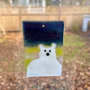 A fused glass hanging ornament featuring a white polar bear with beady eyes. The background on the tile is a midnight blue sky with a lemon coloured aurora borealis.