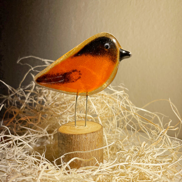 Load image into Gallery viewer, A decorative bird sculpture with a glossy orange body, black head and wing details, and thin metal legs, displayed on a small round wooden base among shredded paper against a warm beige background.