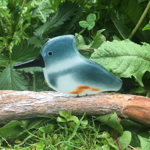 Steel Blue, white and chestnut coloured glass Kingfisher bird ornament perched on driftwood against a background of green foliage.