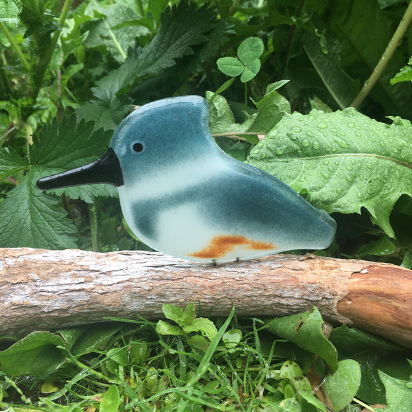Load image into Gallery viewer, Steel Blue, white and chestnut coloured glass Kingfisher bird ornament perched on driftwood against a background of green foliage.