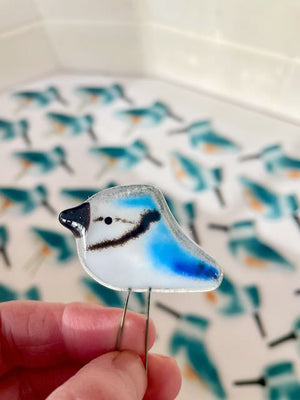A tiny blue and white glass blue jay is being held in the finger tips. In the background are glass kingfishers in a kiln.
