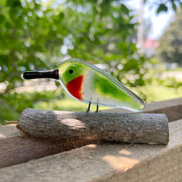 Load image into Gallery viewer, Glass hummingbird figurine with green body and red chest on a wooden log outdoors