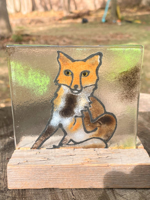 Reverse side of a fused glass panel featuring a fox illustration on a wooden stand , against a natural background