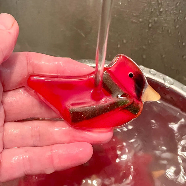 Load image into Gallery viewer, A red glass bird wearing a green glass scarf is being held in the hand and washed under a stream of water.