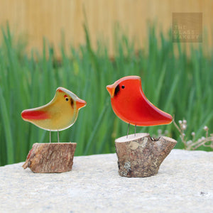 two glass bird ornaments perch on logs on top of a rock. The birds are a male and female.