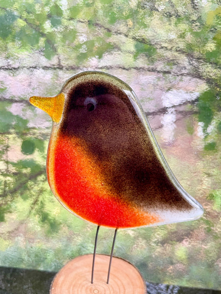 Load image into Gallery viewer, A decorative bird figurine with a glossy brown, orange, and red body, yellow beak, and thin black wire legs, displayed on a small round wooden base against a blurred green outdoor background.