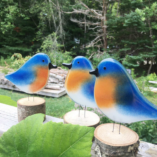 Group of three blue and tan glass Bluebird ornaments in the foreground, with woodland in the background