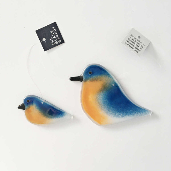 Load image into Gallery viewer, A large and small glass bluebird pair on a white background. The logo for The Glass Bakery is attached to the monofilament hanging from the birds.