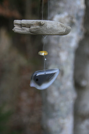 Small grey and white glass bird hanging from driftwood. A yellow glass bead is strung between the bird ornament and the driftwood.