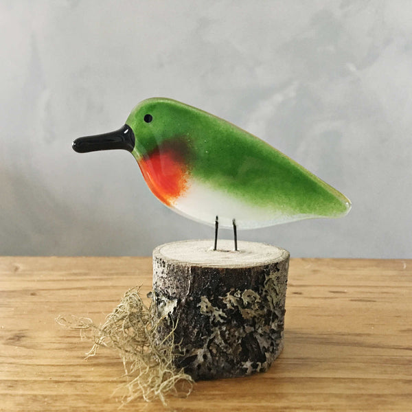 Load image into Gallery viewer, A fused glass red and green ruby throated hummingbird sits on a log. The log is on a table.