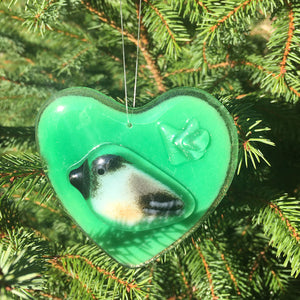 A Green Fused Glass Heart with Chickadee hangs in front of a Christmas Tree