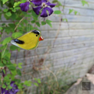 A handcrafted fused glass hanging bird ornament designed to resemble an American Goldfinch, with bright yellow body and black wings, set against a blurred background of flowers.