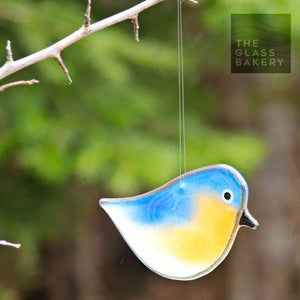 orange, blue and white glass hanging eastern bluebird hangs from a branch, with a tree in the background