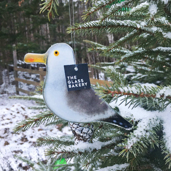 Load image into Gallery viewer, A grey, white and black glass seagull hanging bird hangs from a snowy Christmas tree.