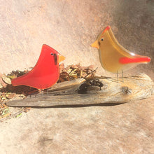 Load image into Gallery viewer, Two fused glass cardinals, one red male and one an amber female, are perched on driftwood. The background is a weathered metal. 
