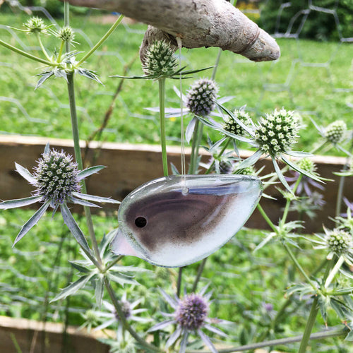 Small grey and white glass bird with pink beak dangles from driftwood. In the background are some thistles and a farm fence