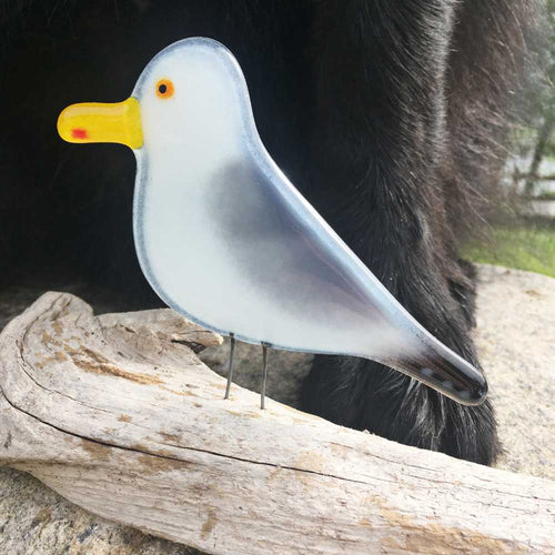 Glass seagull perched on driftwood with a black dog's legs in the background.
