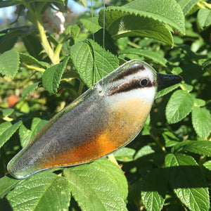 Small glass hanging bird with grey wings, a stripey black and white face, black beak and rusty chest. A close up photograph of the glass bird hanging in front of a rose bush.
