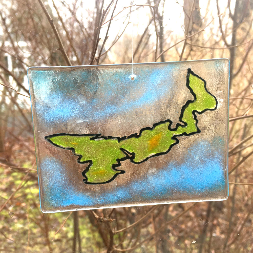 A rectangular glass sun catcher featuring the shape of Prince Edward Island in green and outlined in black. The background glass is blue to represent the sea.