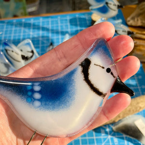 A fused glass blue jay is held in the palm of a hand. In the background are smaller fused glass birds.