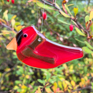 A Red glass Cardinal Hanging Bird is wearing a green glass scarf. The bird hangs in front of a bush with green berries.