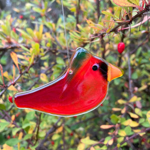 A Red glass Cardinal Hanging Bird is wearing a green glass hat. The bird hangs in front of a bush with green berries.